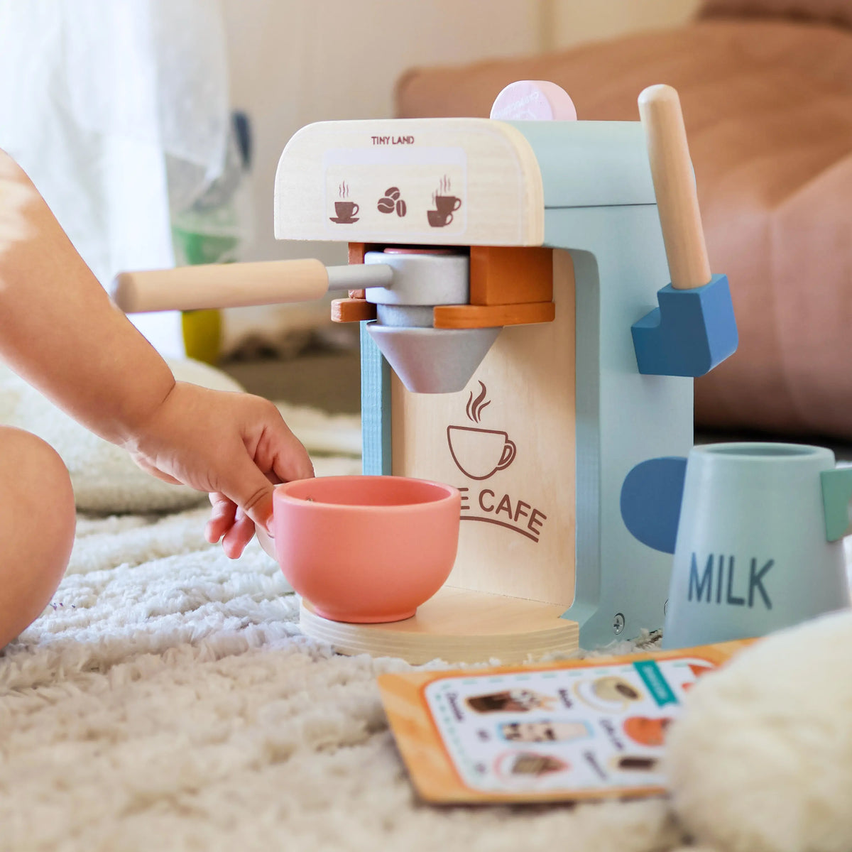 A child’s hand reaches for a pink cup in front of the Tiny Land® Smart Storage Kitchen Cafe Set, featuring a blue milk jug and accessory card on a carpet—perfect for imaginative pretend play from Tiny Land.