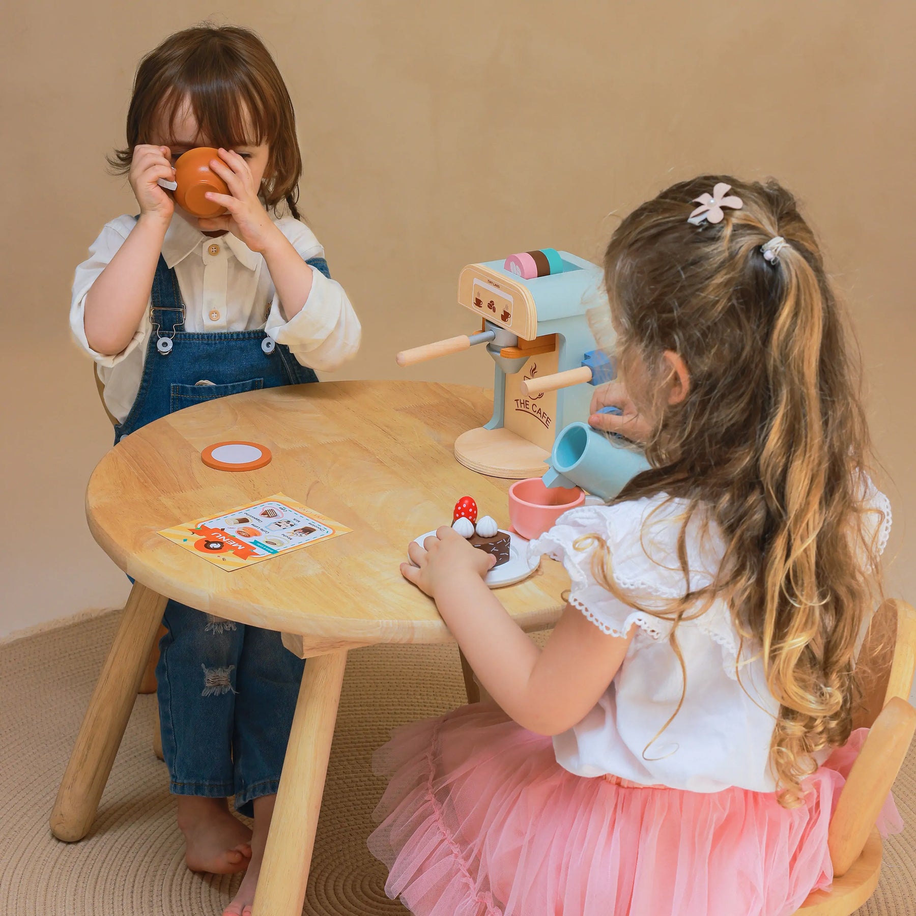 Two children play at a round wooden table with the Tiny Land® Play Kitchen Accessories Children’s Coffee Maker, sipping from toy cups and sharing desserts—enjoying their Tiny Land wooden kids play coffee maker set together.