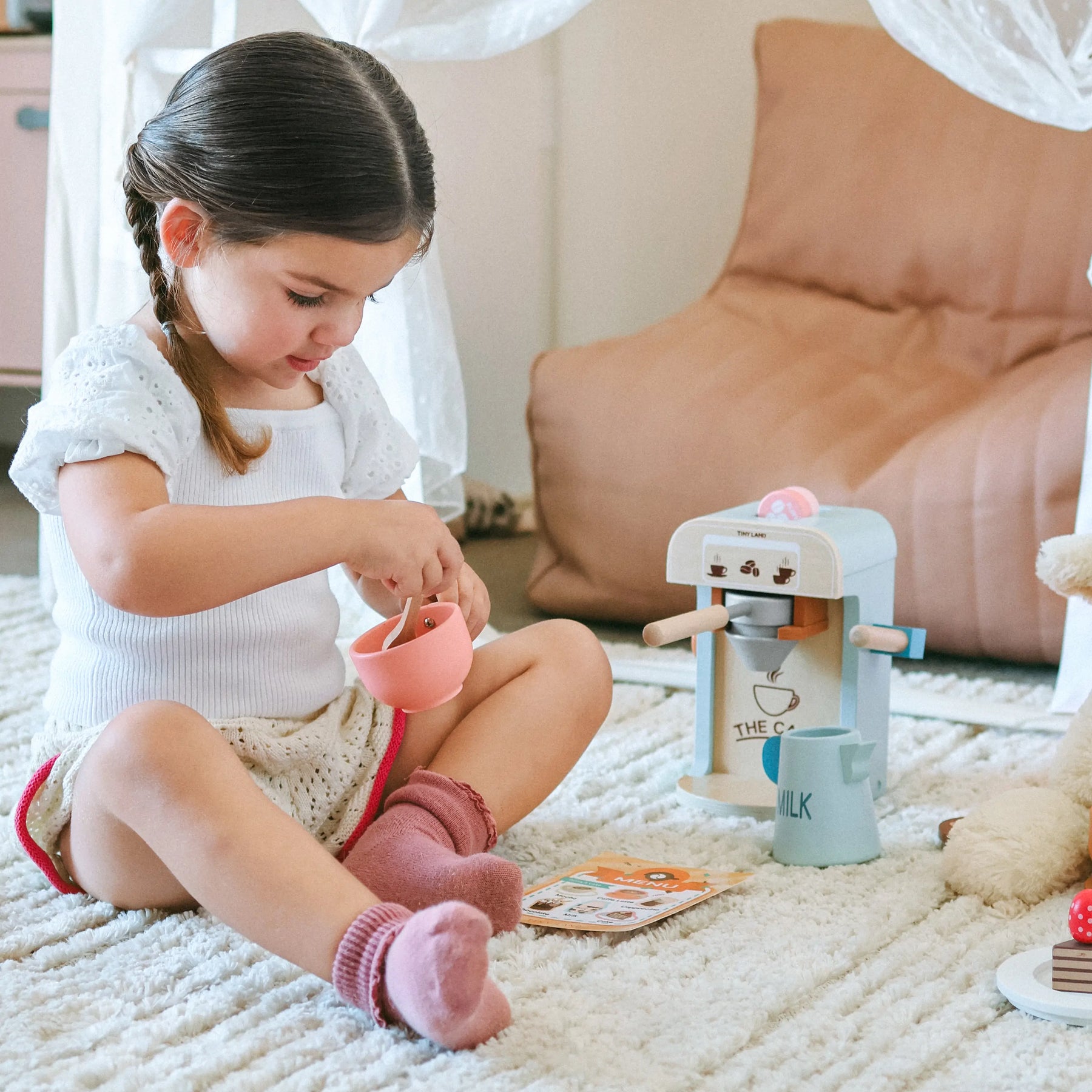 A young girl with brown hair plays on a soft rug with the Tiny Land® Wooden Kids Play Coffee Maker Set, wearing a white outfit and pink socks. She is focused on her imaginative play with kids kitchen toys by Tiny Land in a cozy indoor setting.