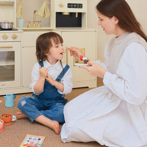 A woman in a white dress sits on the floor, happily feeding a young boy in overalls as they play with Tiny Land® Play Kitchen Accessories – Children’s Coffee Maker, surrounded by wooden kids play items, both engaged and enjoying their time.