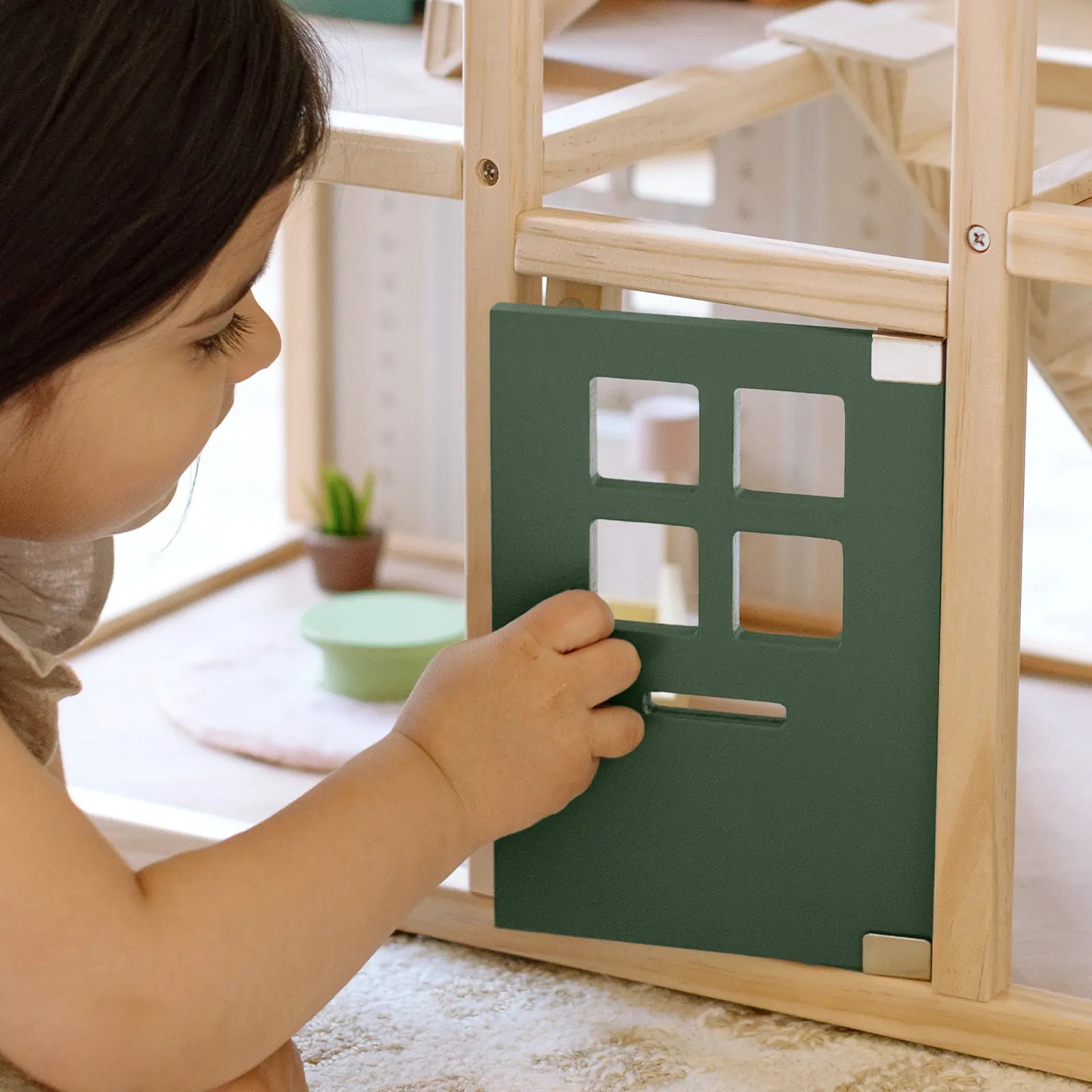 A young child opens the forest green door of the Tiny Land® Sweetwood Evergreen Dollhouse. Inside this furnished Tiny Land dollhouse are miniature items like a small plant and dishes. Dolls not included.|green