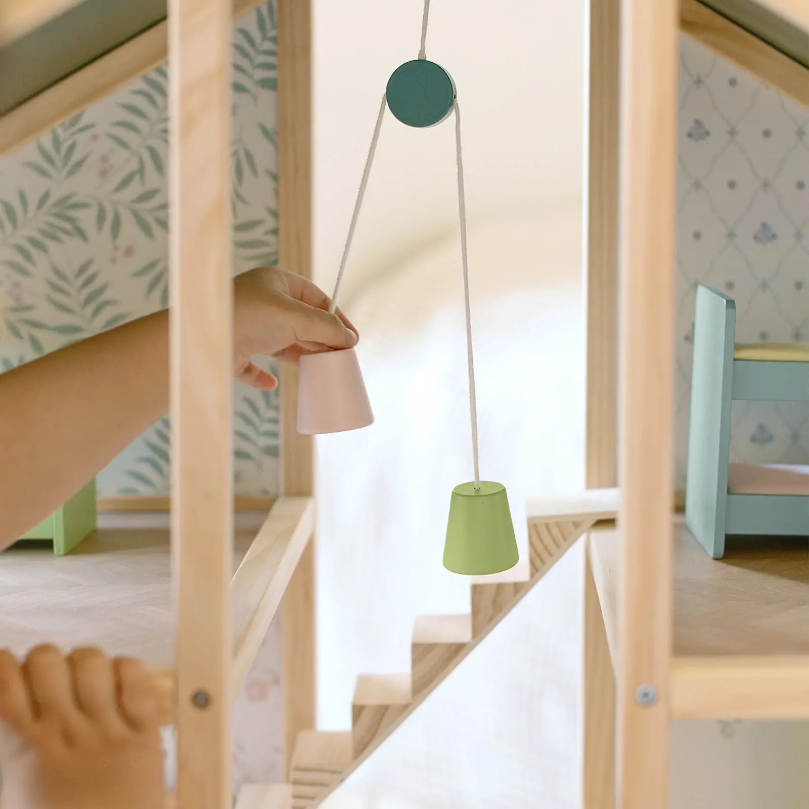 A child operates a small wooden pulley with pink and green buckets inside the Tiny Land® Sweetwood Evergreen Dollhouse by Tiny Land, featuring wallpapered walls and miniature furniture (dolls not included).|green