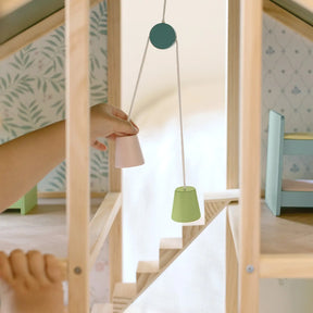 A child operates a small wooden pulley with pink and green buckets inside the Tiny Land® Sweetwood Evergreen Dollhouse by Tiny Land, featuring wallpapered walls and miniature furniture (dolls not included).|green