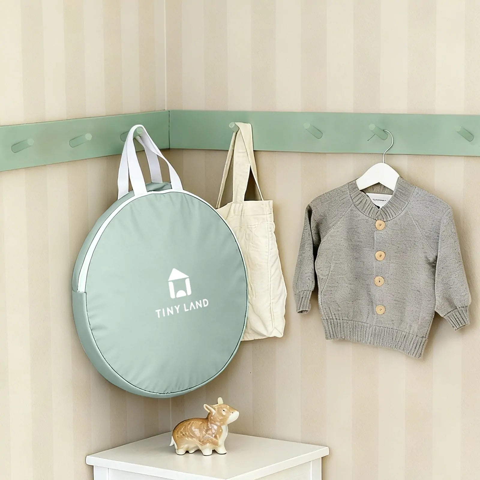 A soft green round bag labeled Tiny Land, a beige tote, and a gray knit sweater hang above a white shelf with a ceramic cow—charming decor for cozy indoor adventures with the Tiny Land® Tiny Sprout Castle Tent.|Green