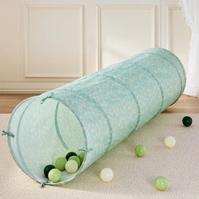 A cheerful indoor playground scene features the Tiny Land® PlayNest Climbing Ball Pit Set with Tunnel by Tiny Land, showcasing a light blue tunnel adorned with a white floral pattern resting on a beige carpet. Green, yellow, and white plastic balls are scattered inside and outside the tunnel, while the PlayNest climbing blocks contribute to the fun-filled atmosphere. In the background, a cream wall and curtain complete the setting.|Blue