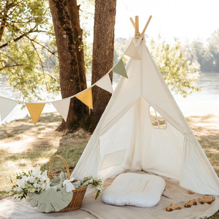 The Tiny Land® Teepee Tent for Kids with Mat by Tiny Land is set up outdoors on a blanket near trees and a lake. The white fabric play tent, featuring a cozy floor mat inside, is decorated with a basket of white flowers and a string of yellow and green pennant flags.