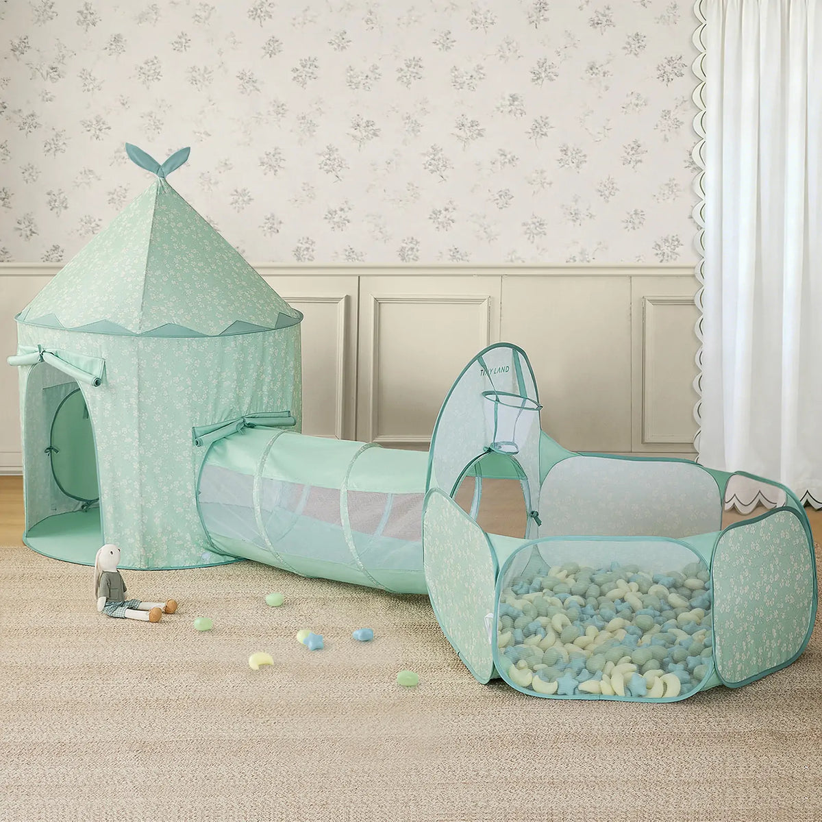 The Tiny Land® Green Adventure 3-in-1 Play Tunnel & Fun-Shapes Ocean Ball Set features a pastel green tunnel connecting to a ball pit filled with soft balls, all set in an elegant, softly lit room with floral wallpaper.