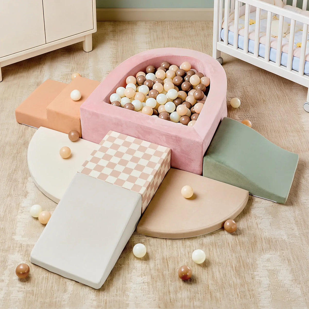 The Tiny Land® PlayNest Climbing & Ball Pit Set by Tiny Land features colorful foam climbing blocks, ramps, and a pink foam ball pit filled with pastel balls, arranged on a beige rug near a crib and dresser in the nursery. This soft indoor playground also includes loose balls scattered around the play area.|Pink