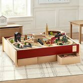 The Tiny Land® 1920 Railway Adventure Train Table from Target features interactive wooden tracks, buildings, and bridges on a red-trimmed play table with storage drawers in a sunlit, carpeted room with wooden furniture.