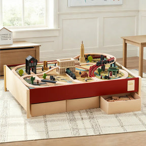 The Tiny Land® 1920 Railway Adventure Train Table from Target features interactive wooden tracks, buildings, and bridges on a red-trimmed play table with storage drawers in a sunlit, carpeted room with wooden furniture.