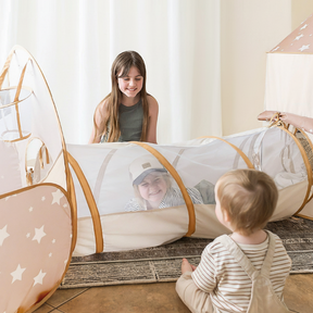 Three children play happily indoors with the Tiny Land® Discovery 5-in-1 Play Tunnel by Tiny Land. One smiles inside, another sits at the entrance, and an older child kneels nearby, all engaged with the beige peachskin polyester kids tunnel.