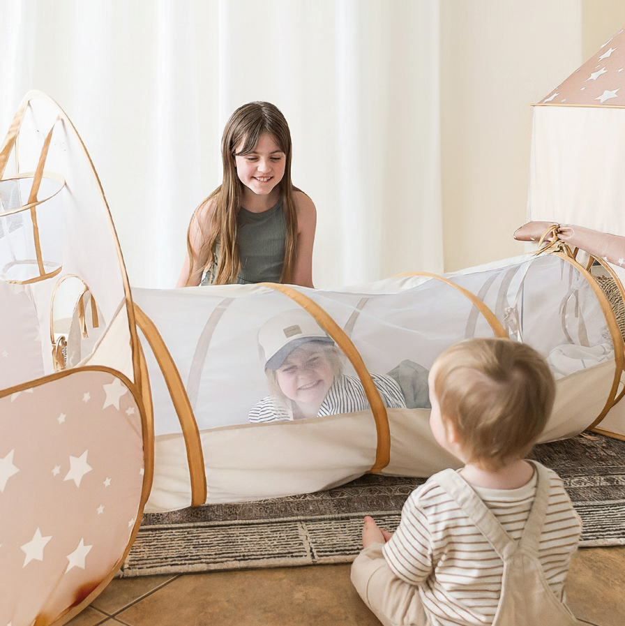 Three children play happily indoors with the Tiny Land® Discovery 5-in-1 Play Tunnel by Tiny Land. One smiles inside, another sits at the entrance, and an older child kneels nearby, all engaged with the beige peachskin polyester kids tunnel.