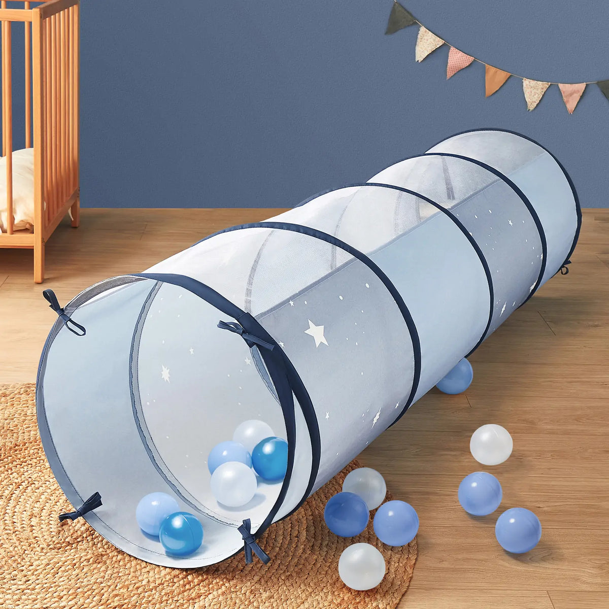 The Tiny Land® Adventure Play Tunnel - Blue by Tiny Land, featuring blue mesh and navy trim, rests on a wooden floor surrounded by blue and white balls in a playfully decorated kids' room with a crib, wall decor, and rug.|blue