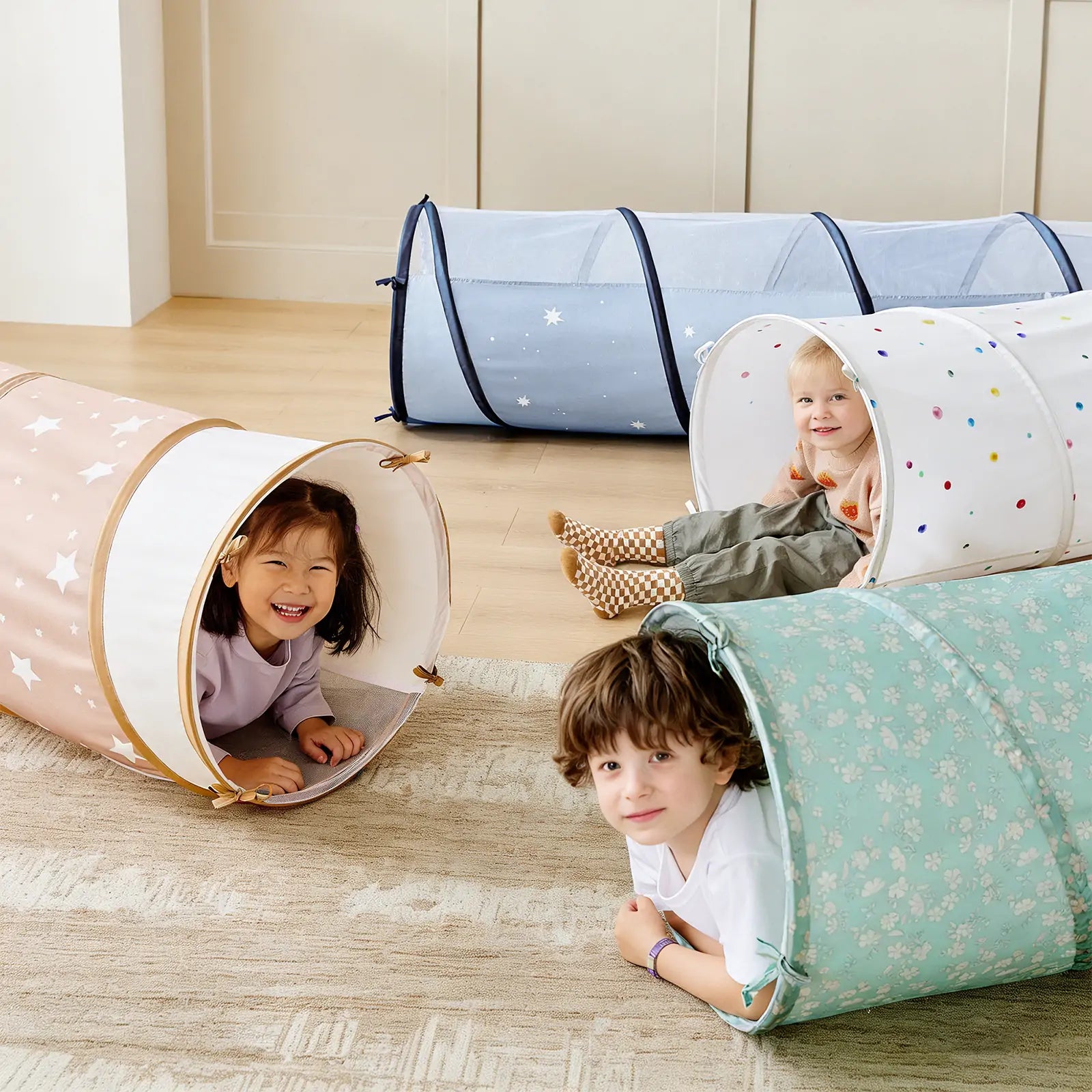 Three children smile as they crawl through the Tiny Land® Adventure Play Tunnel by Tiny Land, which features colorful patterns like stars, polka dots, and floral designs on a wooden floor inside a room. This portable play tunnel adds extra fun and flexibility for both indoor and outdoor adventures.
