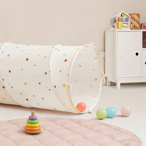 A children's playroom featuring the Tiny Land® Adventure Play Tunnel—Colorful Polka Dots, pastel toy balls, a rainbow ring stacker, a padded pink play mat, a white cabinet, and a colorful toy house on top of the cabinet.