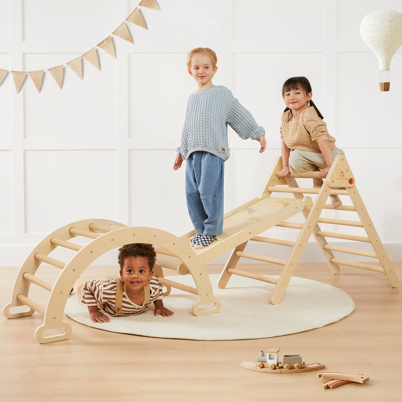 Three children enjoy the Tiny Land Safe Explorers Playset—one stands atop the natural wood climbing set, another climbs, and a third crawls beneath. The Urban Zoo Playmat and fun decor complete the bright, cozy playroom scene.|natural wood color