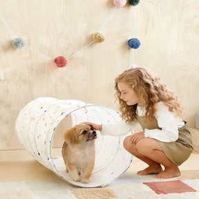 A young girl with curly hair pets a small brown and white puppy inside the Tiny Land® Adventure Play Tunnel - Colorful Polka Dots by Tiny Land, in a room with light wood walls and colorful pom-pom garlands.