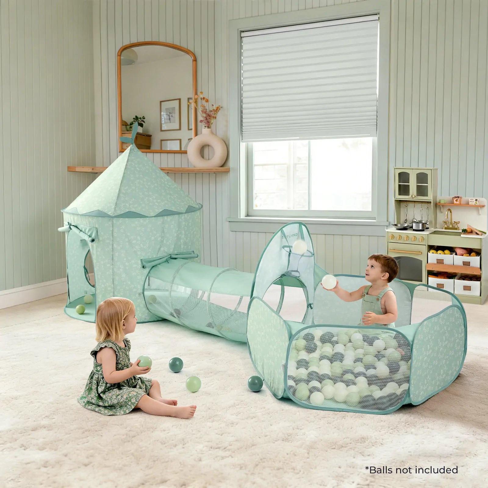 Two children play with the Tiny Land® Adventure 3-in-1 Play Tunnel & Ocean Ball Set - Green, featuring a tent, tunnel, and ball pit. The room has light walls, a window, shelves with toys, and soft balls scattered on the carpet.