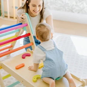 A smiling woman watches a baby in blue overalls climbing on the Tiny Land® 7-in-1 Rainbow Pikler Triangle Set by Tiny Land, featuring colorful bars and a reversible ramp in a bright, cheerful room.|rainbow color