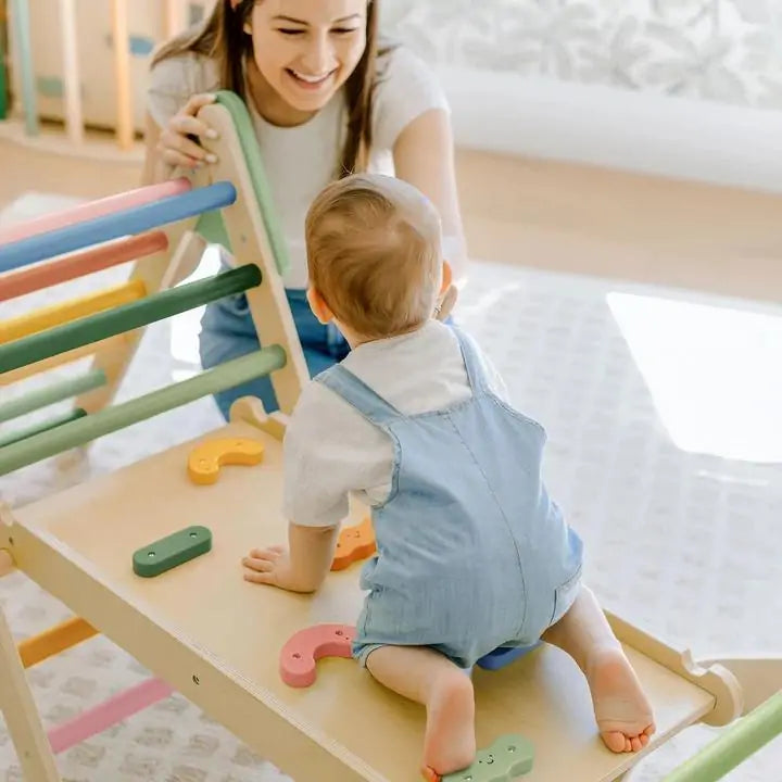 A baby in blue overalls climbs the Tiny Land® 7-in-1 Morandi Pikler Triangle Set by Tiny Land, using colorful handles as an encouraging adult smiles nearby in a bright, child-friendly room.|morandi color