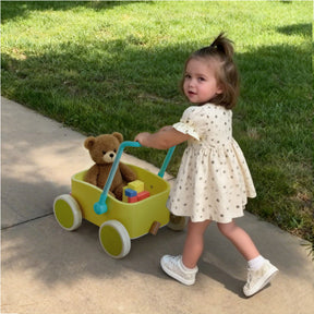 A young girl in a polka-dot dress pushes the Tiny Land® 3-in-1 Wooden Baby Walker Wagon - Spring Color, carrying a teddy bear and blocks along the sidewalk, with green grass behind her.