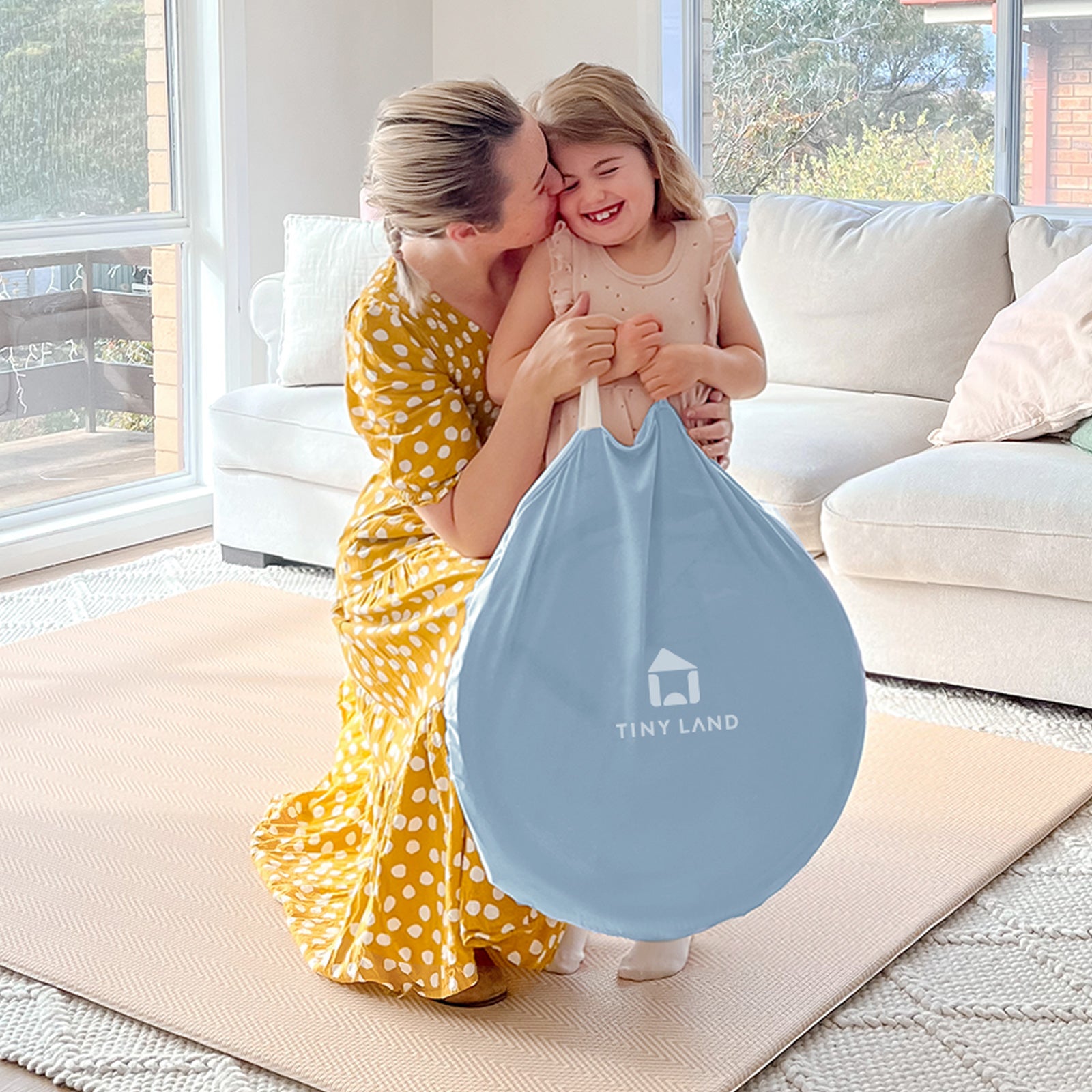 A woman in a yellow polka dot dress hugs a smiling young girl holding a Tiny Land® Kids Tent toy by Tiny Land beside an indoor tent for toddlers in a bright living room with a white sofa and large windows.|Blue