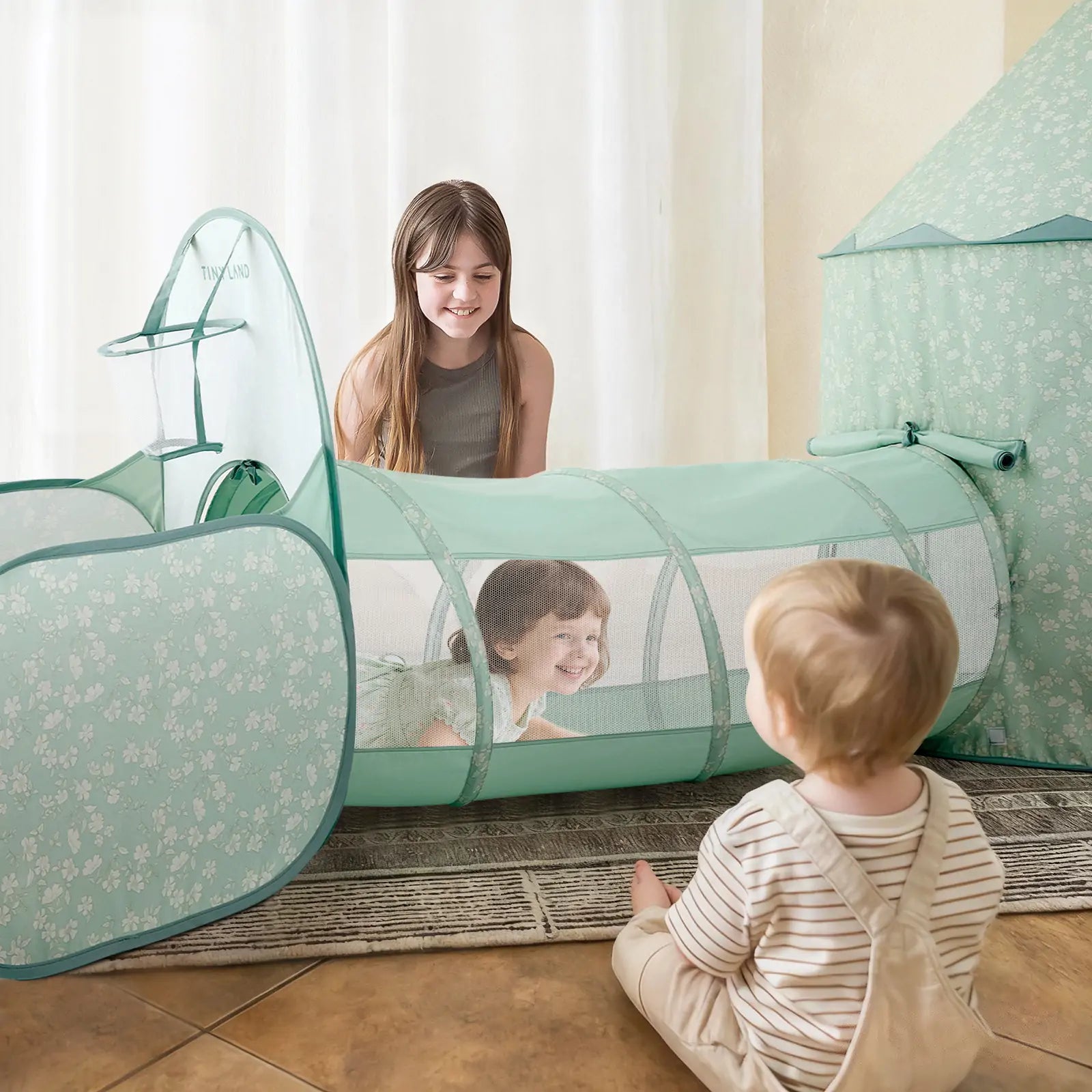 Three children enjoy sensory play indoors with the Tiny Land® Green Adventure 3-in-1 Play Tunnel & Fun-Shapes Ocean Ball Set—one smiles inside the tunnel, one sits at the entrance, and another kneels nearby, all engaged in imaginative fun.