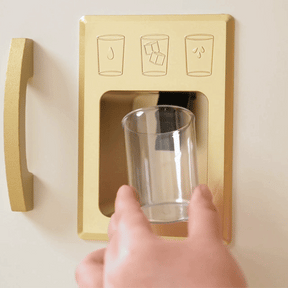 A hand holds a glass under the golden ice dispenser of the Tiny Land® PopJoy Kitchen & Fridge Set as three ice cubes drop in—ideal for kids' pretend play fun by Tiny Land.