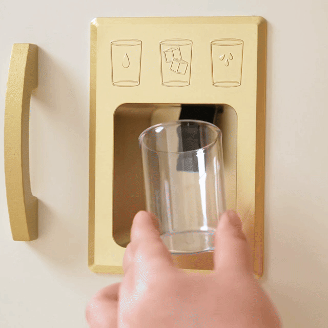 A hand holds a glass under the golden ice dispenser of the Tiny Land® PopJoy Kitchen & Fridge Set as three ice cubes drop in—ideal for kids' pretend play fun by Tiny Land.