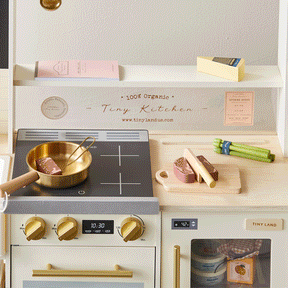 The Tiny Land® BistroSwitch Kitchen & Fridge Set by Tiny Land is a miniature play kitchen for kids, featuring a stovetop, pan, cutting board, toy food items, and small kitchen accessories neatly arranged on light-colored shelves and counters.