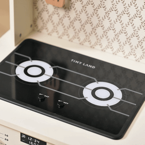 A close-up of the Tiny Land® Farm-to-Table Play Set’s toy stovetop shows two black knobs beneath burners with white and gray circular designs—ideal for pretend play in this interactive kitchen from Tiny Land.