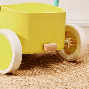 A close-up of the Tiny Land® 3-in-1 Wooden Baby Walker Wagon in Spring Color, featuring yellow panels, white-rimmed wheels, a wooden side peg, adjustable speed control, and a turquoise handle on a natural fiber rug.