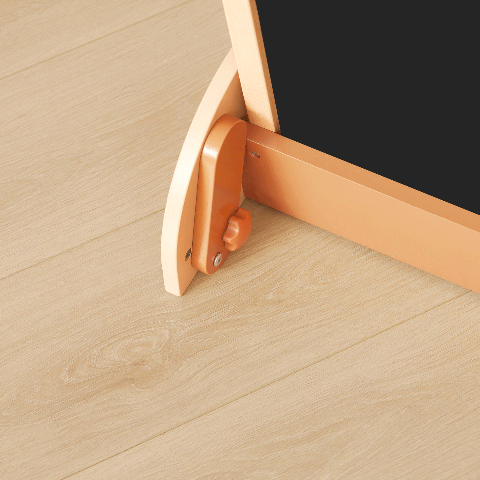 Close-up view of the base of the Tiny Land® Double-Sided Arched Magnet Chalkboard & Art Kit, showcasing a rounded light-wood leg, an orange adjustment knob, and a coordinating orange support bar—all by Tiny Land—resting on a light-colored wooden floor.