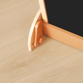 Close-up view of the base of the Tiny Land® Double-Sided Arched Magnet Chalkboard & Art Kit, showcasing a rounded light-wood leg, an orange adjustment knob, and a coordinating orange support bar—all by Tiny Land—resting on a light-colored wooden floor.