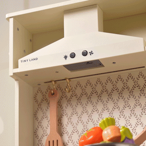A close-up of Tiny Land's Farm-to-Table Play Set features an interactive kitchen range hood with control buttons, a hanging wooden utensil, patterned wallpaper, and organic toy vegetables—perfect for imaginative play.