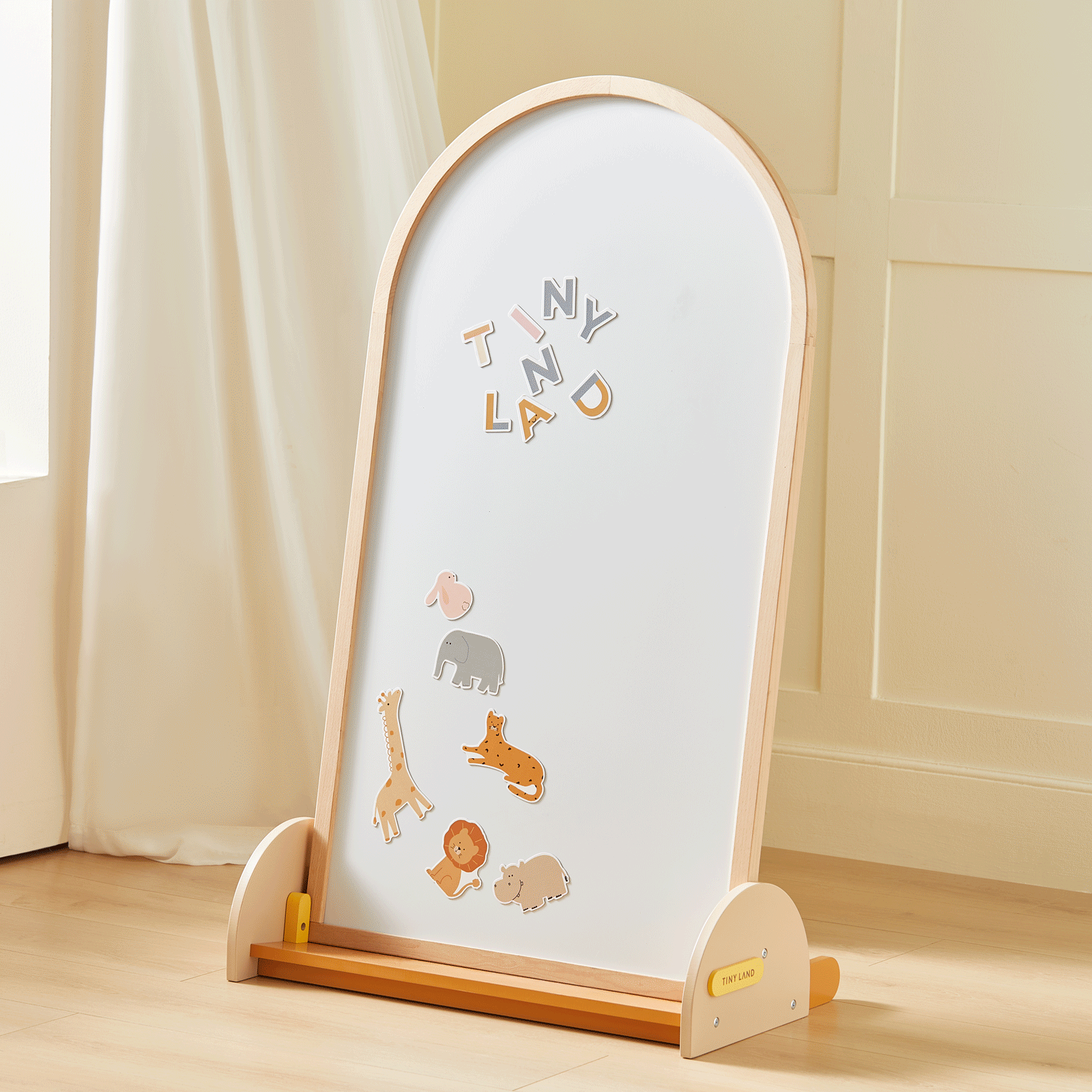 A wooden-framed Tiny Land® Double-Sided Arched Magnet Chalkboard & Art Kit stands on a light floor, showcasing colorful animal magnets and letter magnets that spell out TINY LAND. This versatile easel from Tiny Land is perfect for creative play, with cream curtains and a paneled wall visible in the background.