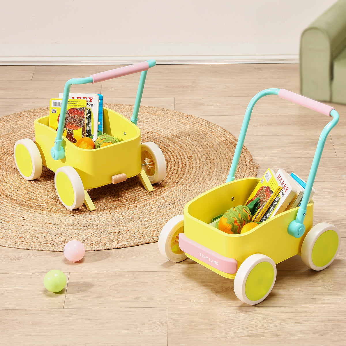 Two Tiny Land® 3-in-1 Wooden Baby Walker Wagons in yellow with blue handles and white wheels sit on a round woven rug, each holding children’s books and toy vegetables. Colorful balls are scattered on the light wooden floor nearby.