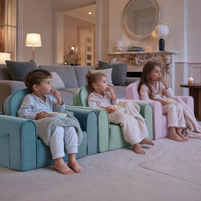 Three young children in pajamas sit on colorful Tiny Land® GentleSnug Kids Sofa Chairs and a toddler sofa in a cozy living room, enjoying popcorn while watching something offscreen, with a larger sofa, fireplace, and kids nap mat visible in the background.|Light Green