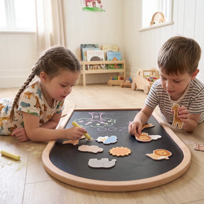 Two young children play with animal-shaped pieces and draw on the Tiny Land® Double-Sided Arched Magnet Chalkboard in a bright, cozy room with wooden shelves and toys in the background.