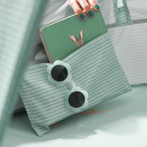 A hand with teal-painted nails holds a tablet showing a rabbit, partly tucked in the Tiny Land® 3-in-1 Baby Beach Tent bag. Flower-shaped sunglasses sit on the bag, and sand is sprinkled over the mint surface.