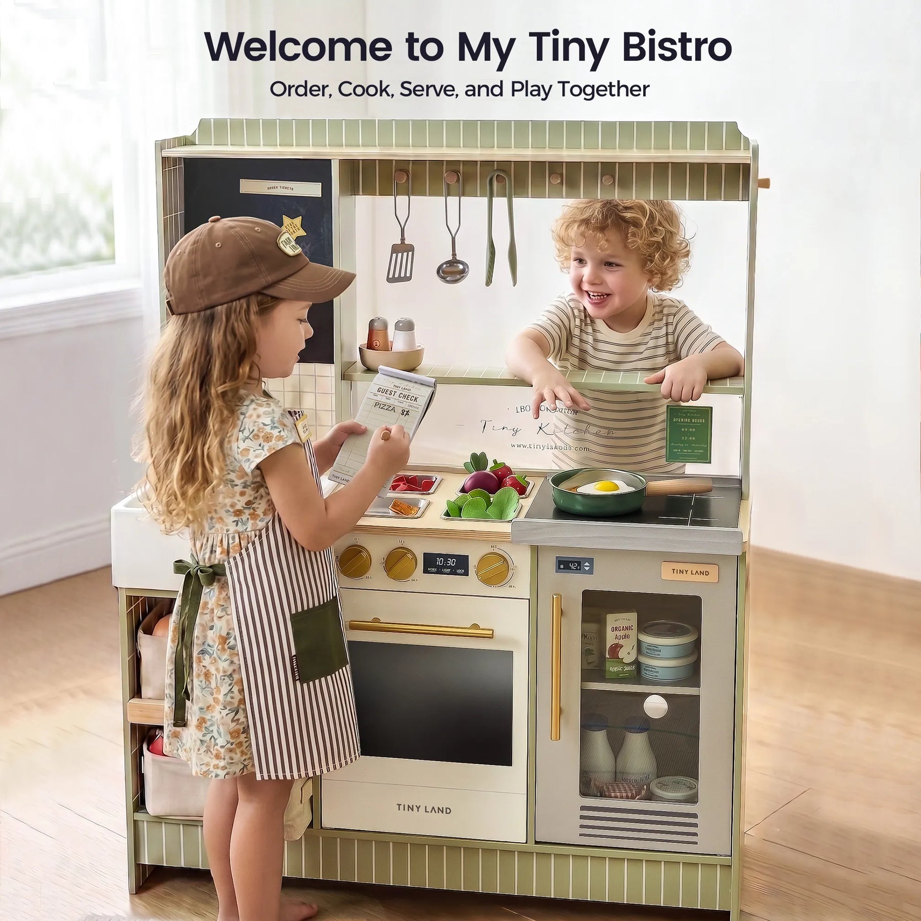 Two young children play with the Tiny Land® BistroSwitch Play Kitchen by Tiny Land, pretending to cook and serve food. The girl wears an apron and cap, while the boy stands behind the counter, smiling. A sign reads, "Welcome to My Tiny Bistro.|Green