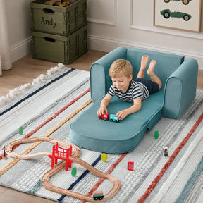 A young boy relaxes on the Tiny Land® GentleSnug Kids Sofa Chair in teal, with its footrest extended, as he plays with toy trains on a wooden train set atop a striped rug. Green storage boxes and car-themed decor from Tiny Land are visible in the background.|Blue