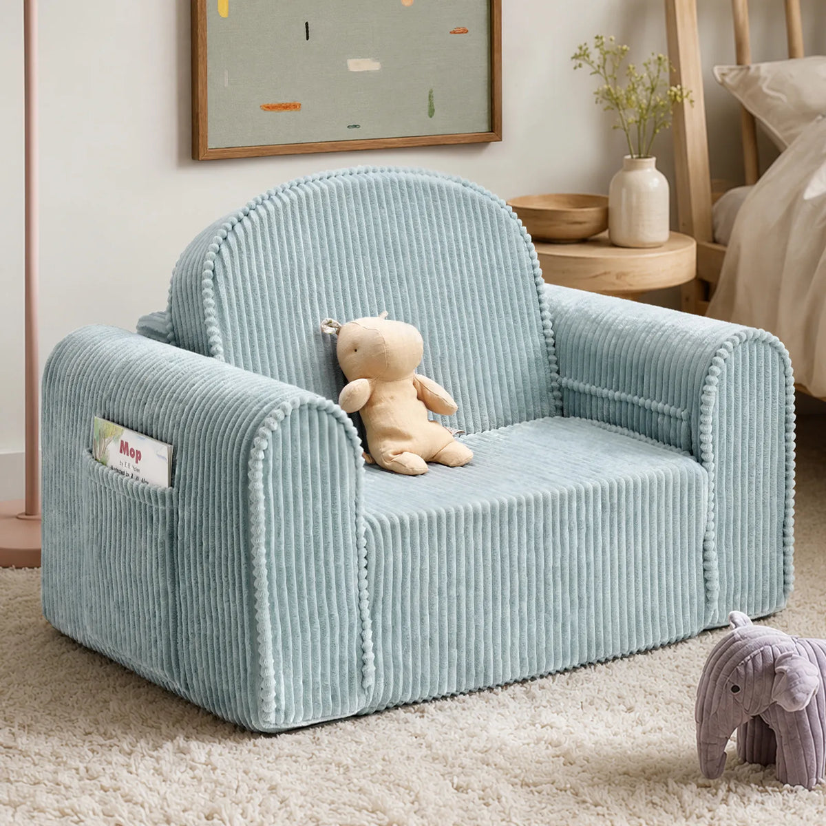 The Tiny Land® GentleSnug Corduroy Kid Chair in light blue serves as a charming kids lounger, adorned with a stuffed bear, and is set in a cozy room featuring a framed art piece, wooden furniture, and a plush elephant toy on a textured rug.|Soft Teal