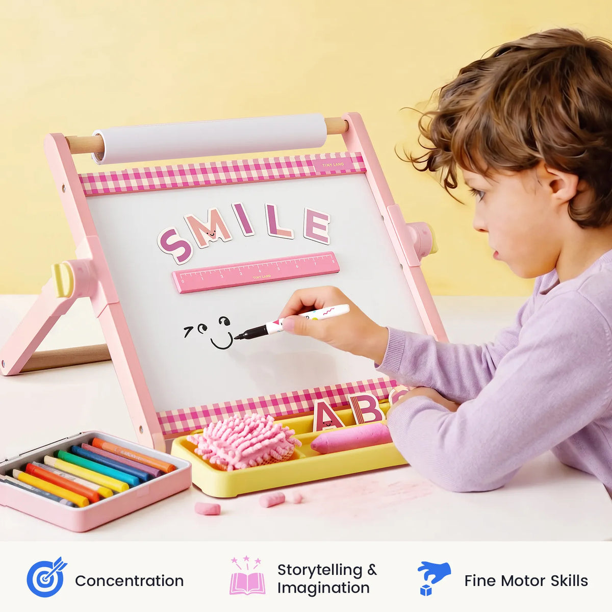 A young child draws a smiling face on the Tiny Land® Double-Sided Magnetic Tabletop Easel, with "SMILE" spelled out in pink letters. Colorful chalk, an eraser, and icons for concentration, imagination, and motor skills are pictured, making this adjustable easel from Tiny Land the perfect kids drawing board for creative play.|Pink