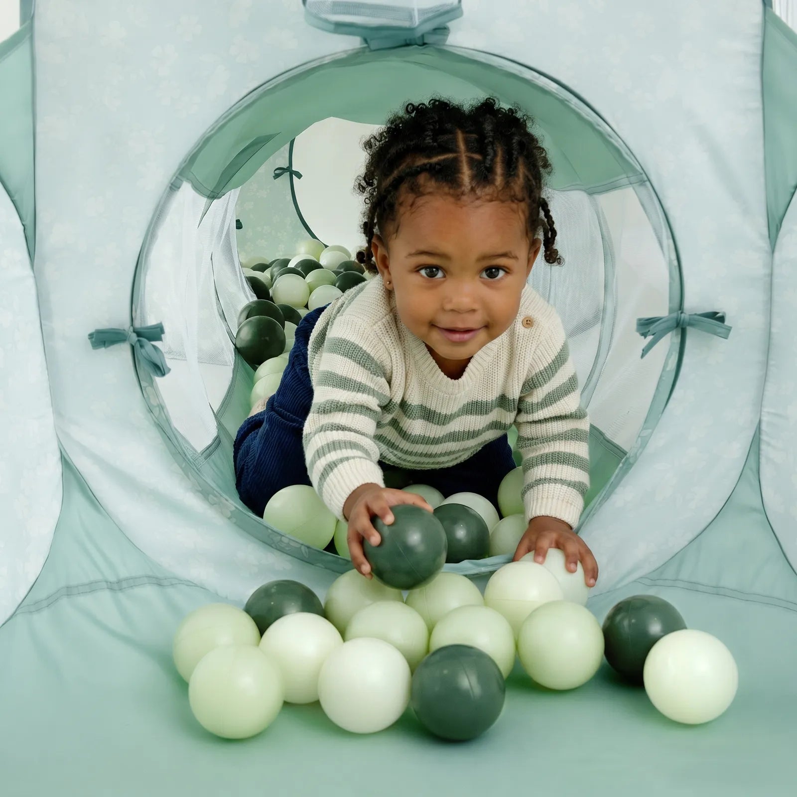 A young child with braided hair crawls through the Tiny Land® Compact Adventure 3-in-1 Play Tunnel, smiling and reaching forward inside a vibrant soft play area.|green