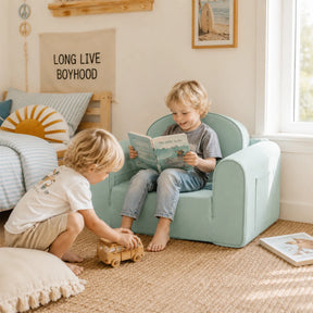 Two young boys play in a sunlit room. One sits in a Tiny Land® GentleSnug Kids Sofa Chair reading a book, while the other plays with a toy car on the floor. The space features playful, beach-themed decor.
