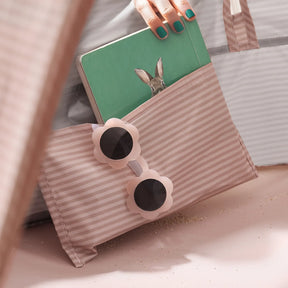 A hand with teal nail polish holds a green book featuring a rabbit, partially tucked in a pink-and-white striped pouch with flower sunglasses—fun accessories for lounging under the Tiny Land® 3-in-1 Baby Beach Tent by Tiny Land.
