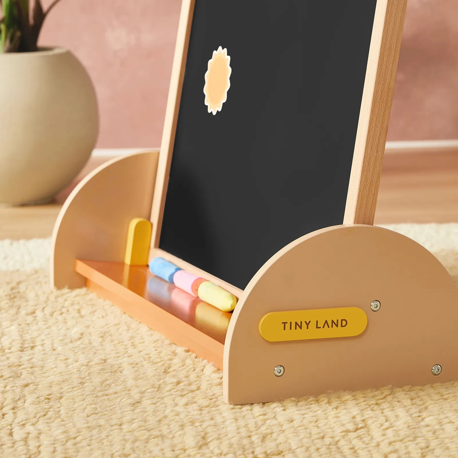 The Tiny Land® Double-Sided Arched Magnet Chalkboard, featuring whiteboard and chalkboard surfaces with colored chalk, rests on a soft beige rug. A potted plant can be seen in the background.