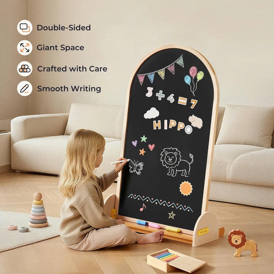 A young child draws with chalk on the Tiny Land® Double-Sided Arched Magnet Chalkboard, featuring math, balloons, and animal doodles. Wooden Tiny Land lion toy and stacked rings rest nearby on the wooden floor.