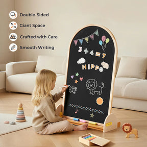 A young child draws with chalk on the Tiny Land® Double-Sided Arched Magnet Chalkboard, featuring math, balloons, and animal doodles. Wooden Tiny Land lion toy and stacked rings rest nearby on the wooden floor.